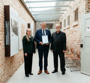 Drei Personen in hellem Gang, Ausstellung der Gedenkstätte mit Infotafeln an den Wänden. Die drei blicken freundlich, Gordon Schnieder hält seine Auszeichnung als Schirmherr in den Händen.