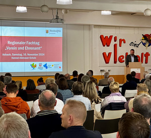Chef der Staatskanzlei, Dr. Fedor Ruhose, steht vor einem sitzenden Publikum am Podium und hält eine Rede. Im Hintergrund eine Leinwand, auf die Regionaler Fachtag "Verein und Ehrenamt" projiziert wird.