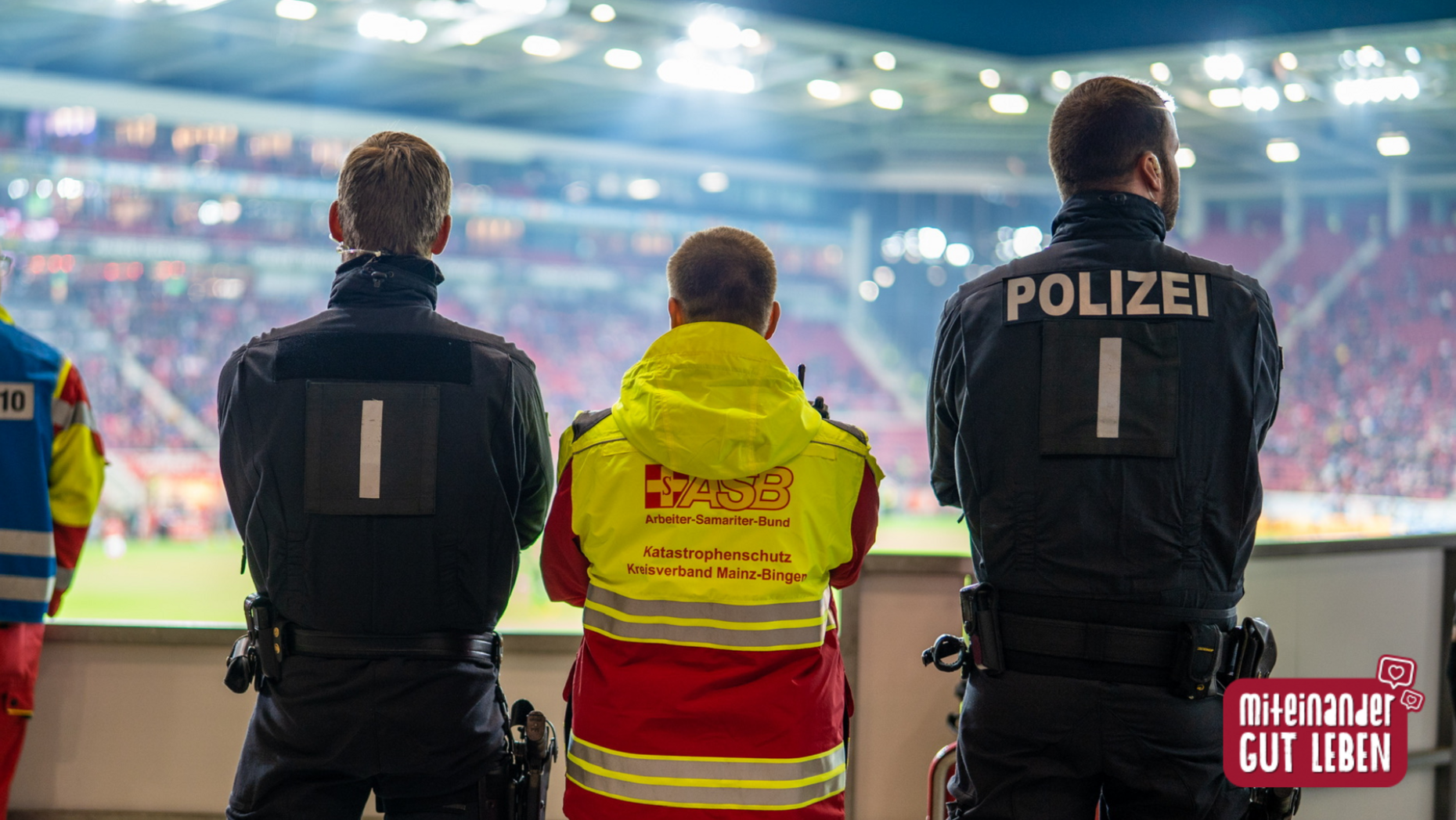 Zwei Polizisten und eine Rettungskraft vom ASB stehen vor einer Stadionkulisse.