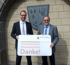 Der Chef der Staatskanzlei, Staatssekretär Clemens Hoch, und Verbandsbürgermeister Gerd Heilmann halten eine Ehrenamtskarte in großem Format; Bild: VG Vordereifel