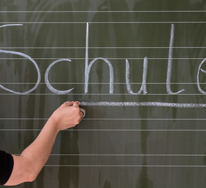 Ein Kind schreibt mit Kreide das Wort Schule an die Tafel.